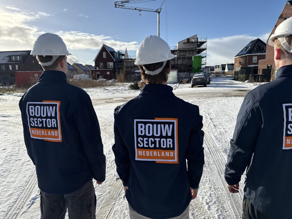 Drie vakmensen van Bouwsector Nederland in blauwe jassen en witte helmen bij een nieuwbouwstraat met hijskraan